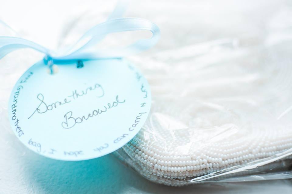 A round blue tag with "Something Borrowed" written in script adorns a white beaded handbag for a bride on her wedding day.