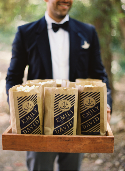Groomsman in tux holds wooden tray with paper popcorn bags that read "Emily and David"